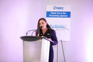 Lora at a podium speaking during the 2025 Diversity and Inclusion Building Science Forum. An IIBEC-branded sign is behind her.