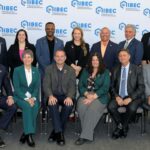 Group of professionals in business attire posing for a group photo at an IIBEC event with a branded backdrop featuring the IIBEC logo.