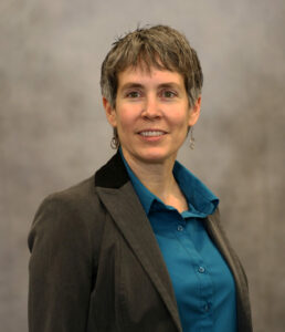 Professional woman in a teal blouse and dark blazer, smiling at the camera, neutral studio backdrop.