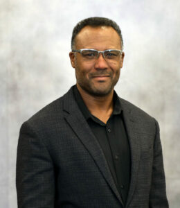 Portrait of a man in a dark blazer and black shirt, wearing glasses, smiling slightly against a neutral backdrop (head-and-shoulders).