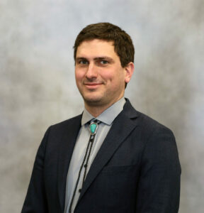 Professional head-and-shoulders portrait of a smiling man in a dark blazer and light gray shirt with a turquoise bolo tie, neutral studio background.