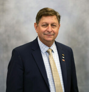 Portrait of a smiling man in a navy suit, light checkered shirt, beige tie, with lapel pins against a gray backdrop.