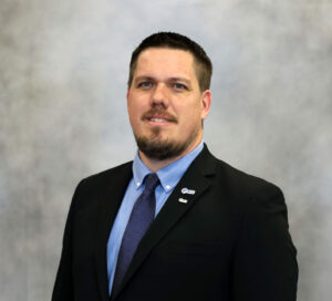 Professional headshot of a man in a dark suit, blue shirt, and patterned blue tie, smiling at the camera with lapel pins visible on the blazer.