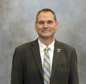 Portrait of a smiling man in a dark suit and plaid yellow-blue tie, against a gray backdrop, with two lapel pins (IBEC and flag).