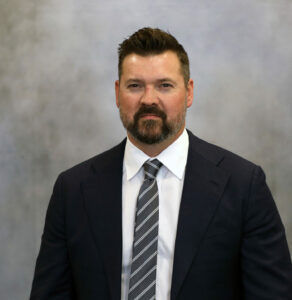 Portrait of a man in a dark suit, white shirt, and striped tie against a gray studio backdrop.