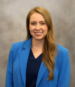 Smiling woman with long blonde-brown hair in a blue blazer and navy top, neutral backdrop.