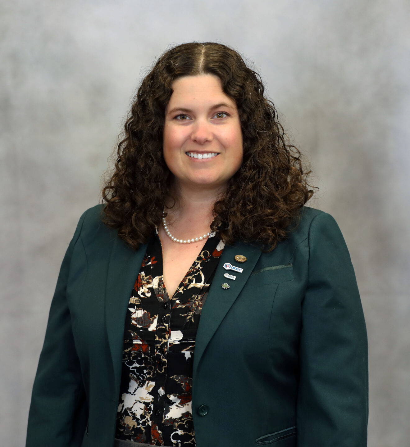Smiling woman with shoulder-length curly hair wearing a teal blazer, pearl necklace, and lapel pins in a professional portrait.