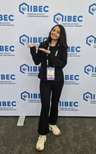 Smiling woman in black suit posing with hands raised in front of an IBEC backdrop at a conference, wearing a name badge.