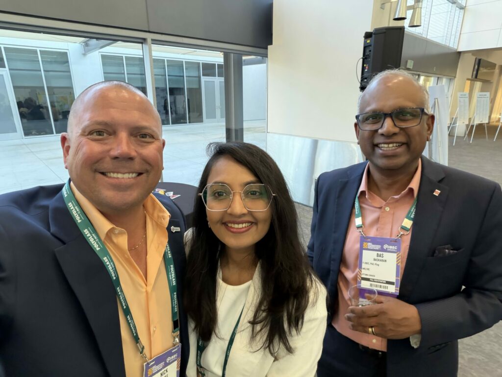 Three professionals in business attire pose for a selfie at a conference, with visible name badges and lanyards in a modern indoor venue.