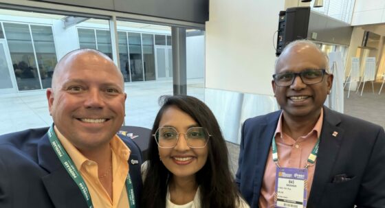 Three professionals in business attire pose for a selfie at a conference, with visible name badges and lanyards in a modern indoor venue.