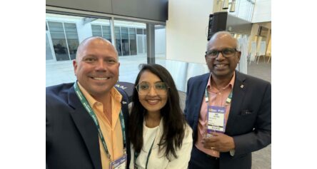 Three professionals in business attire pose for a selfie at a conference, with visible name badges and lanyards in a modern indoor venue.