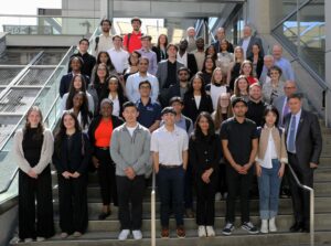 Diverse group of people posed on a wide outdoor staircase in front of a modern glass-and-concrete building for a group photo.