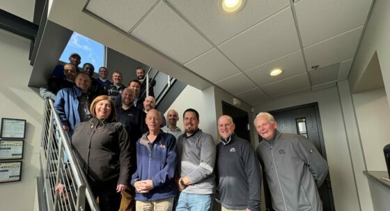 Group of people posing on a stairway indoors, smiling at the camera, wearing jackets and work shirts in a hallway setting.