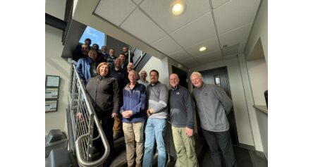 Group of people posing on a stairway indoors, smiling at the camera, wearing jackets and work shirts in a hallway setting.
