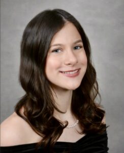 Portrait of a smiling woman with wavy dark hair, wearing a pearl necklace and an off-the-shoulder black top, against a gray studio backdrop.