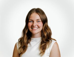 Portrait of a smiling woman with long brown hair wearing a white sleeveless top, against a light background.