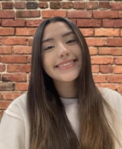 Portrait of a smiling woman with long dark hair wearing a light sweater, standing in front of a brick wall.