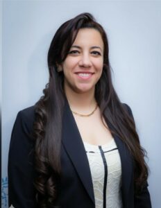 Smiling woman with long dark hair wearing a black blazer against a light blue background.