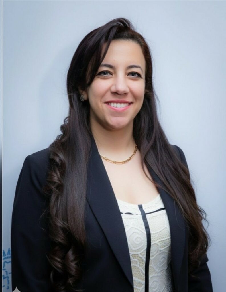 Smiling woman with long dark hair wearing a black blazer against a light blue background.