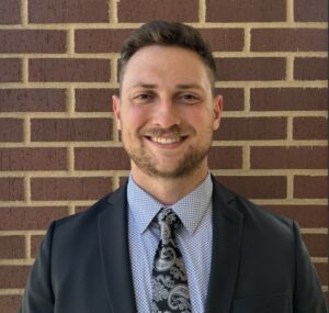 Smiling man in a dark blazer and patterned tie posing against a brick wall.