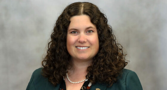 Professional portrait of a smiling woman with curly brown hair, wearing a teal blazer, patterned blouse, and pearl necklace, posed against a neutral backdrop.
