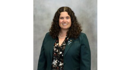 Professional portrait of a smiling woman with curly brown hair, wearing a teal blazer, patterned blouse, and pearl necklace, posed against a neutral backdrop.