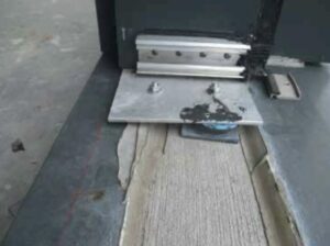 Close-up of a metal floor transition with a sliding track and bolts in a workshop doorway, showing wear and a rubber mat beneath.
